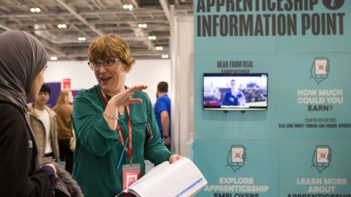 A student is pictured speaking to an exhibitor, on a blue exhibition stand with a large sign saying 'Apprenticeship Information Point'
