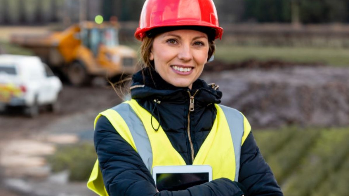 A photo of a young woman, wearing a hard hat and hi-vis jacket, stood in a rural setting holding an ipad